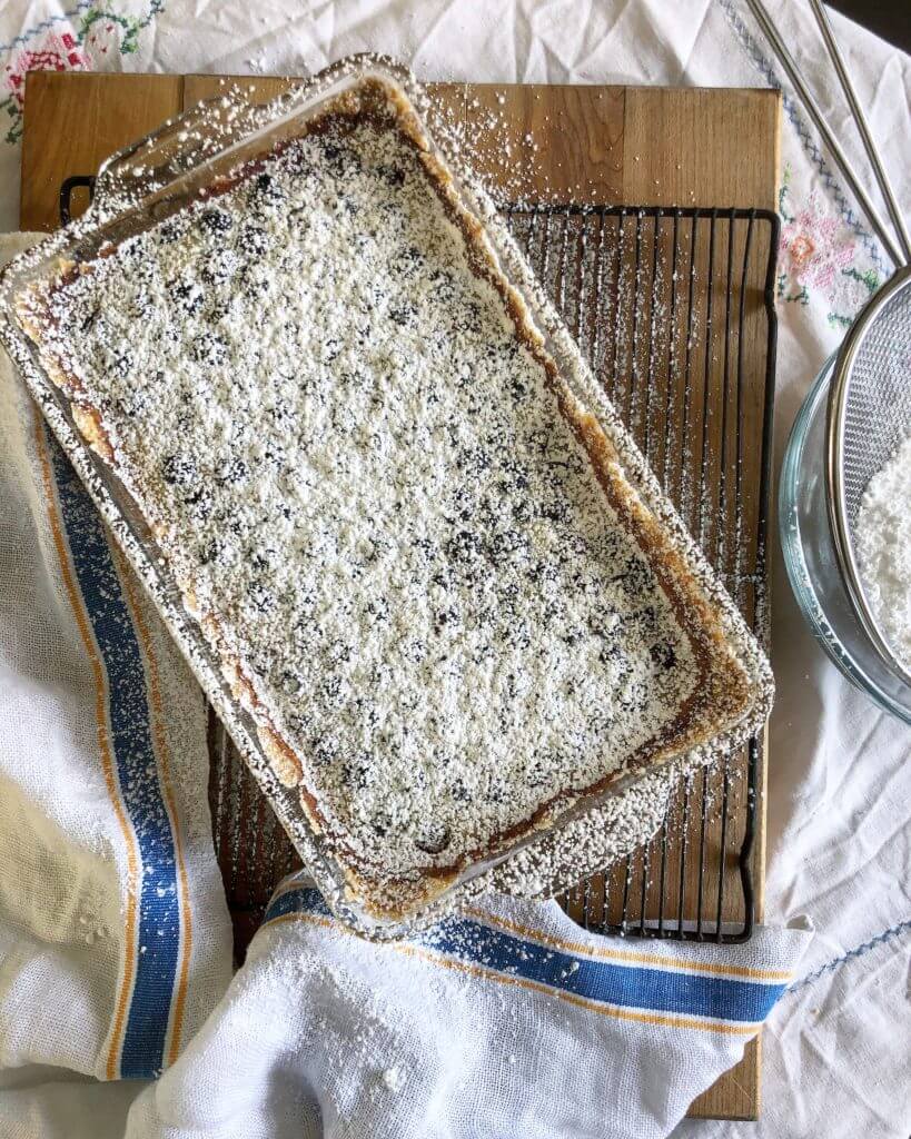 A top view of a baked lemon blueberry dessert in a glass dish, heavily dusted with powdered sugar, resting on a cooling rack with a bowl of extra powdered sugar nearby.