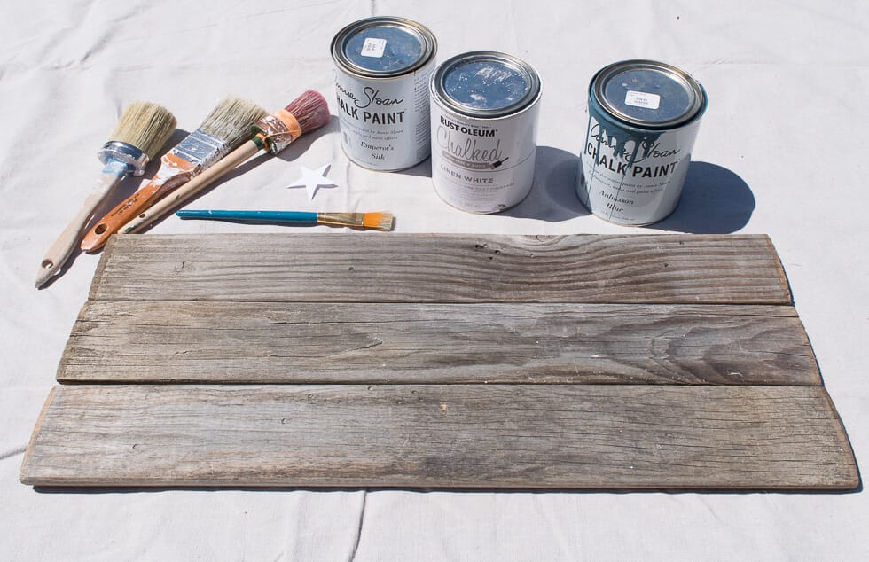 A wooden board, three paint cans labeled as chalk paint, and several paintbrushes are laid out on a light surface.