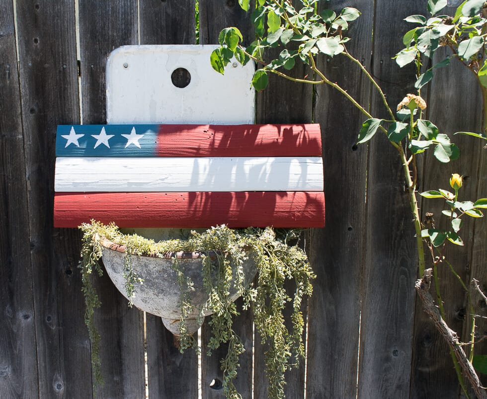 A painted wooden plank resembling the American flag is mounted on a wooden fence, with a planter below it filled with trailing greenery.