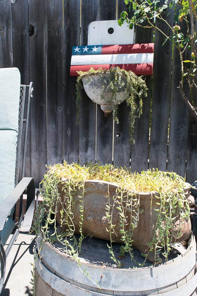 A rustic outdoor setup featuring a painted American flag wooden plank mounted on a fence, with a planter below it and another large, overflowing planter sitting on a barrel nearby.