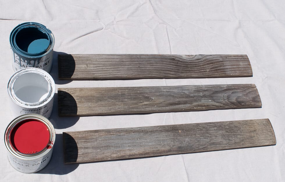 Three paint cans in blue, white, and red are placed beside three unpainted wooden planks on a light surface.