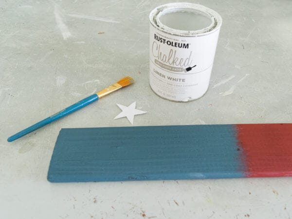 A partially painted wooden plank in blue and red is placed beside a small white star, a paintbrush, and an open can of white chalk paint.