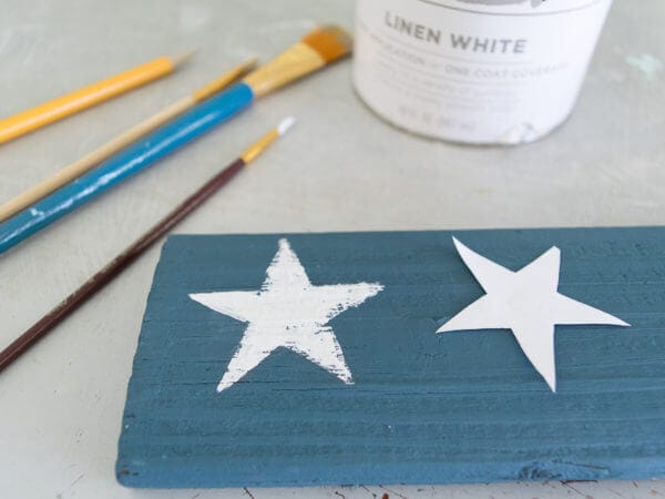 A blue wooden plank with one white painted star and another unpainted star stencil, alongside paintbrushes and a can of white chalk paint.