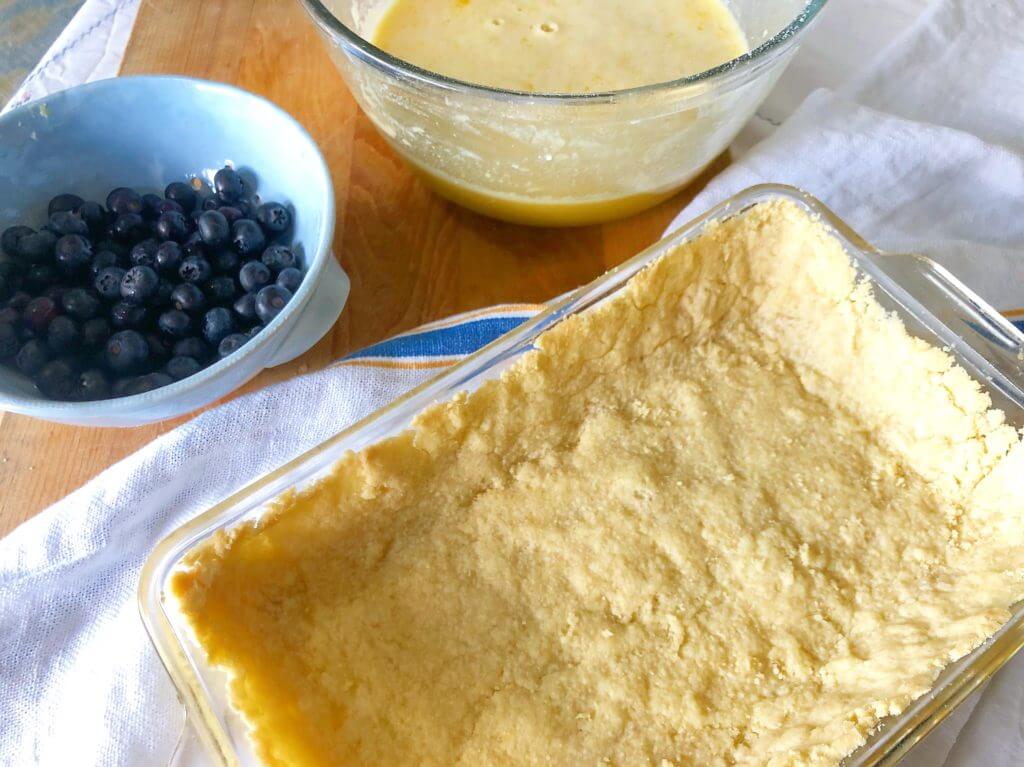 A glass baking dish with an unbaked crust, next to a bowl of blueberries and a mixing bowl containing a lemon filling.