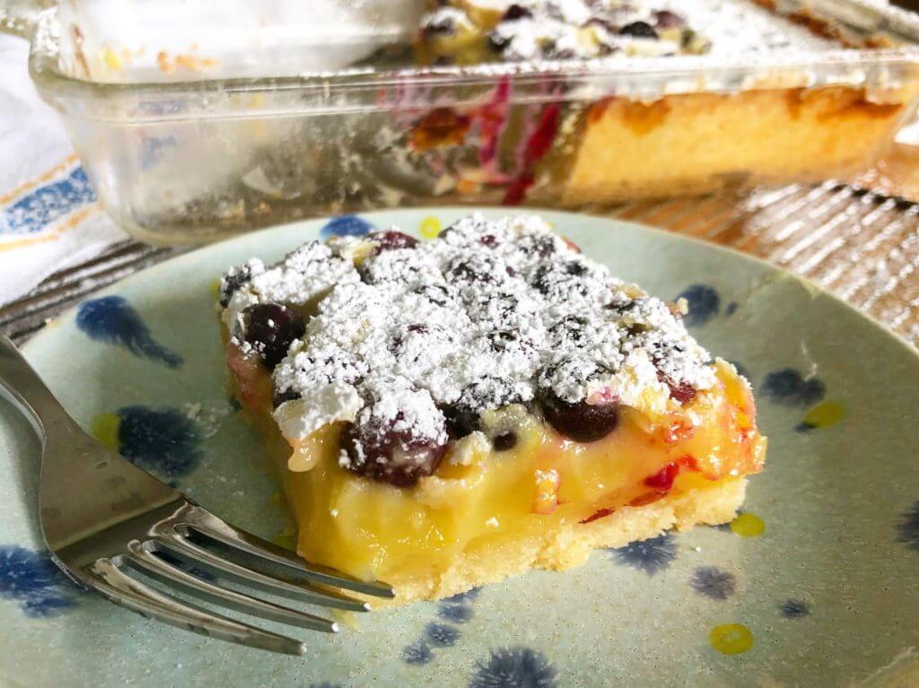 A slice of blueberry lemon bar dusted with powdered sugar on a green plate with a fork.