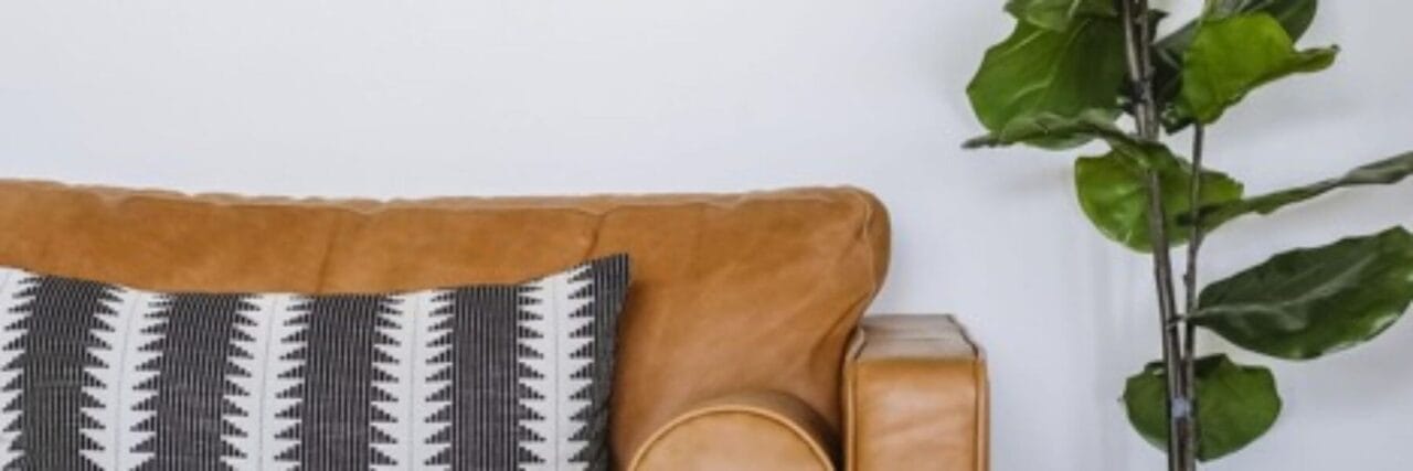 A cozy interior scene featuring a tan leather couch with a black and white patterned pillow, positioned next to an artificial fiddle leaf fig tree.