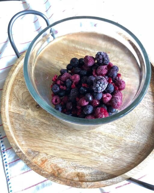 A glass bowl filled with a mixture of frozen berries, including raspberries, blackberries, and blueberries, placed on a round wooden tray.