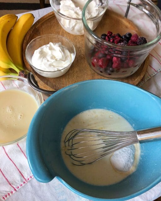 A blue mixing bowl with batter and a whisk, surrounded by ingredients for a dessert and a bowl of custard on a wooden tray.