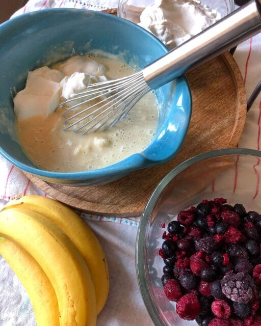 A blue mixing bowl with creamy mixture and a whisk, surrounded by ingredients including bananas and a glass bowl of frozen mixed berries.
