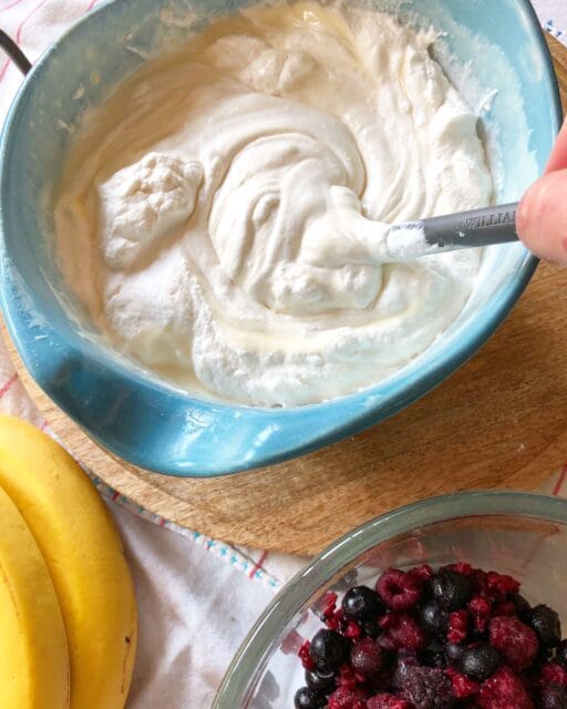 A blue mixing bowl filled with creamy, whipped dessert mixture being stirred with a spoon, with bananas and a glass bowl of frozen mixed berries nearby.