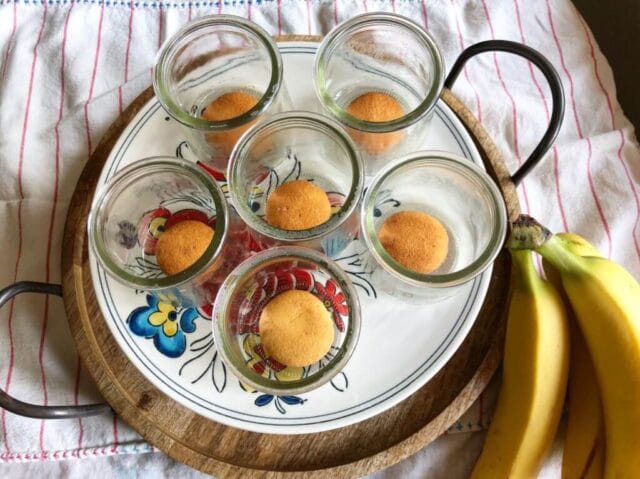 Six small glass jars with vanilla wafers at the bottom, arranged on a round tray with two bananas on the side.