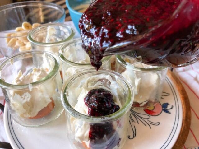 A jar being filled with berry sauce, surrounded by other jars containing layers of vanilla wafers and cream mixture on a round tray.