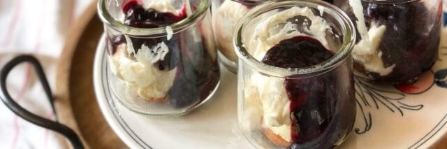 Four glass jars filled with layers of banana pudding, whipped cream, and berry sauce on a decorative round tray.