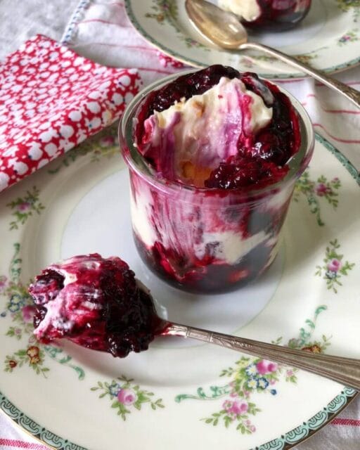 A glass jar filled with berry banana pudding, featuring layers of whipped cream and berry sauce, served on a decorative plate with a spoonful of the dessert beside it.