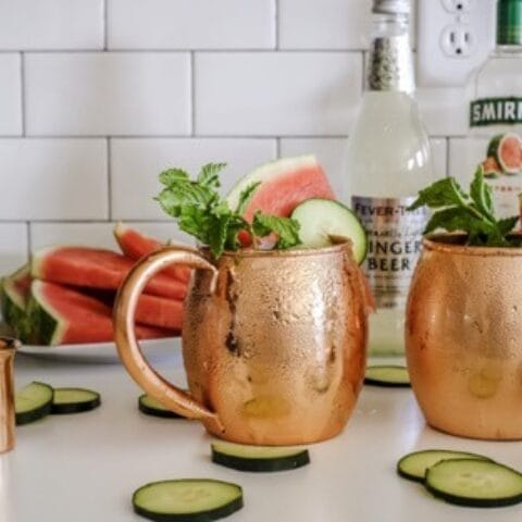 Two copper mugs filled with cucumber and watermelon slices, garnished with mint leaves, with bottles of ginger beer and Smirnoff vodka in the background.