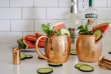Two copper mugs filled with cucumber and watermelon slices, garnished with mint leaves, with bottles of ginger beer and Smirnoff vodka in the background.