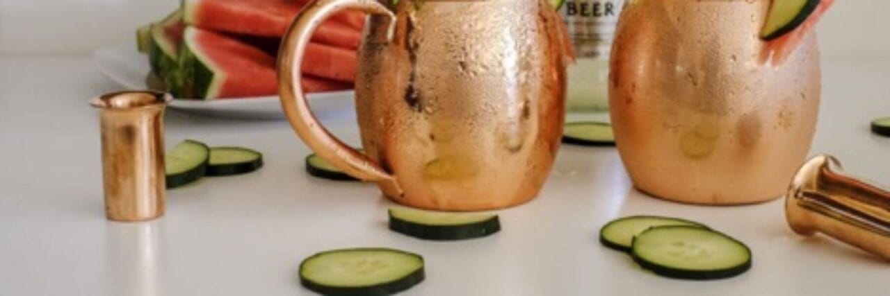 Copper mugs with cucumber slices on a white surface, alongside a plate of watermelon slices in the background.