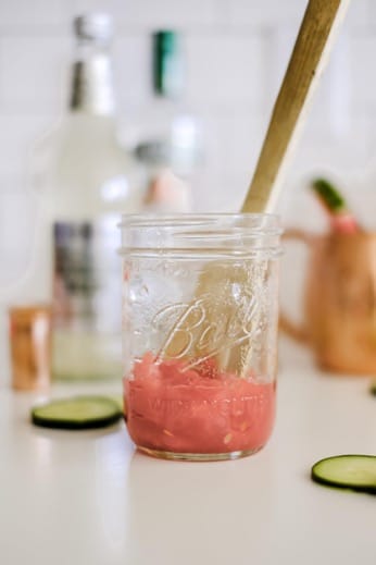 A glass jar with a wooden muddler and mashed watermelon inside, with cucumber slices and a bottle in the background.