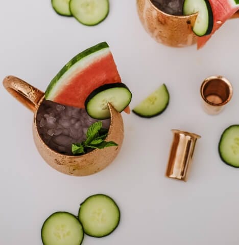 A copper mug filled with crushed ice, garnished with a watermelon slice, cucumber slice, and mint leaves, with more cucumber slices and a small copper cup on a white surface.