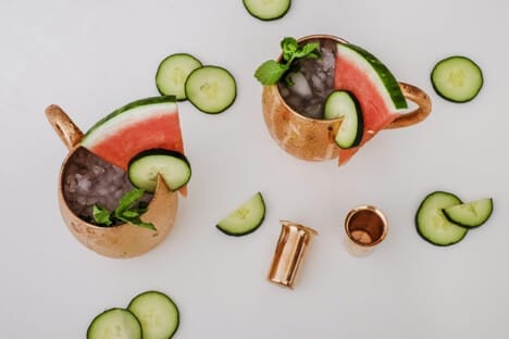 Two copper mugs filled with crushed ice, watermelon slices, cucumber slices, and mint leaves, surrounded by additional cucumber slices on a white surface.