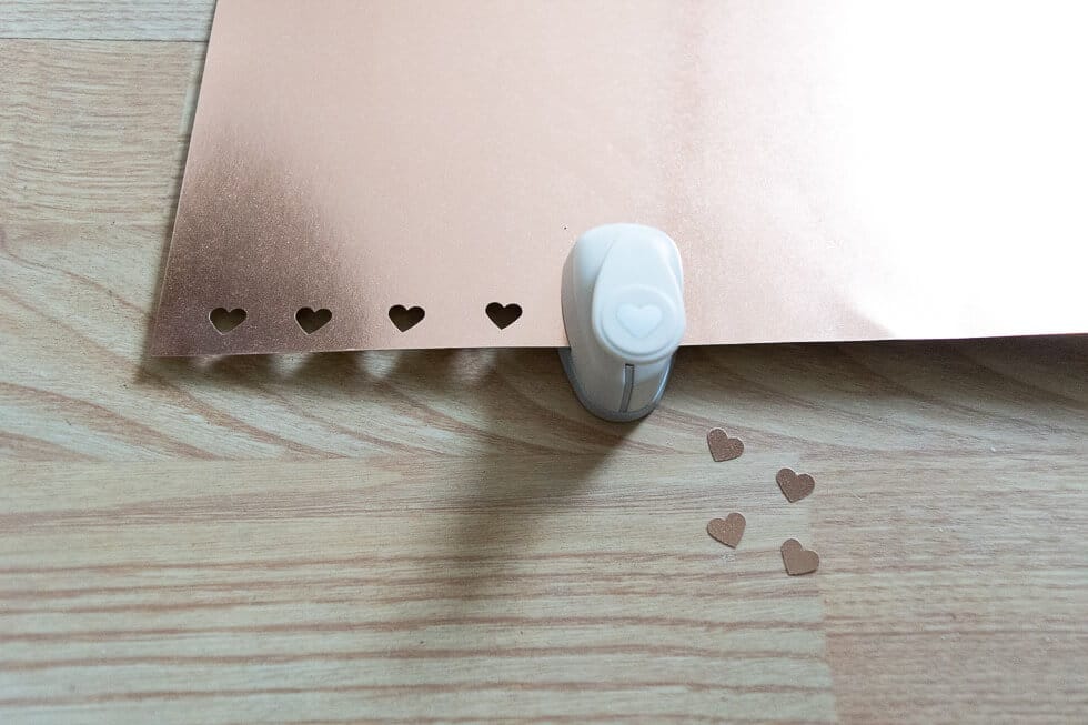 A small heart-shaped punch next to a copper sheet with heart cutouts, with several punched-out heart pieces scattered nearby.