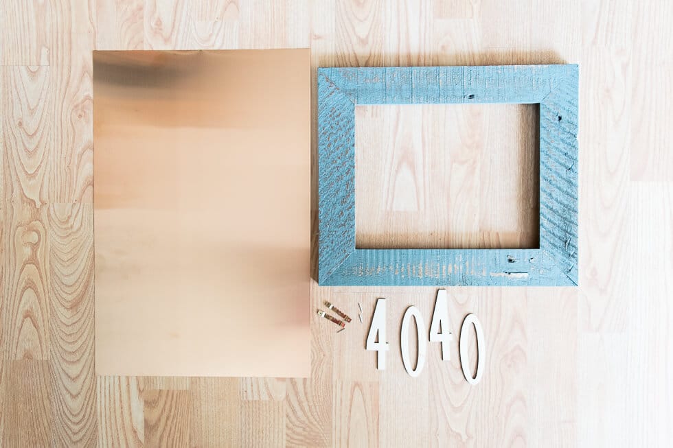 A flat lay of crafting materials on a light wood surface, including a copper sheet, a rustic blue wooden frame, small white numbers "4040," and a few small hanging hooks.