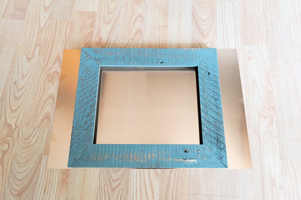 A blue rustic wooden frame placed on top of a copper sheet, both set on a light wood floor. The copper sheet extends beyond the edges of the frame.