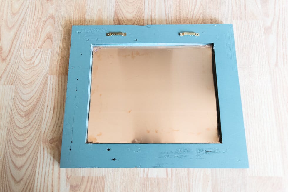 The back of a blue rustic wooden frame, now secured with a copper sheet in the center. The frame has two small hanging hooks attached at the top, and it is placed on a light wood floor.