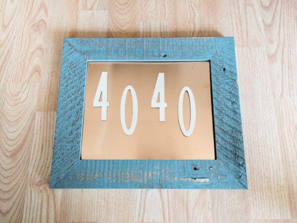 A blue rustic wooden frame with a copper sheet backing, displaying the numbers "4040" in white, arranged in the center. The frame is set on a light wood floor.
