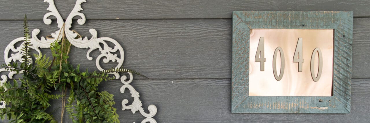 A decorative outdoor scene featuring a house number sign "4040" in a blue rustic wooden frame with a copper backing, mounted on a dark gray wooden wall. To the left, a white ornate wall hanging holds a wreath of green ferns, adding a touch of greenery to the setup.