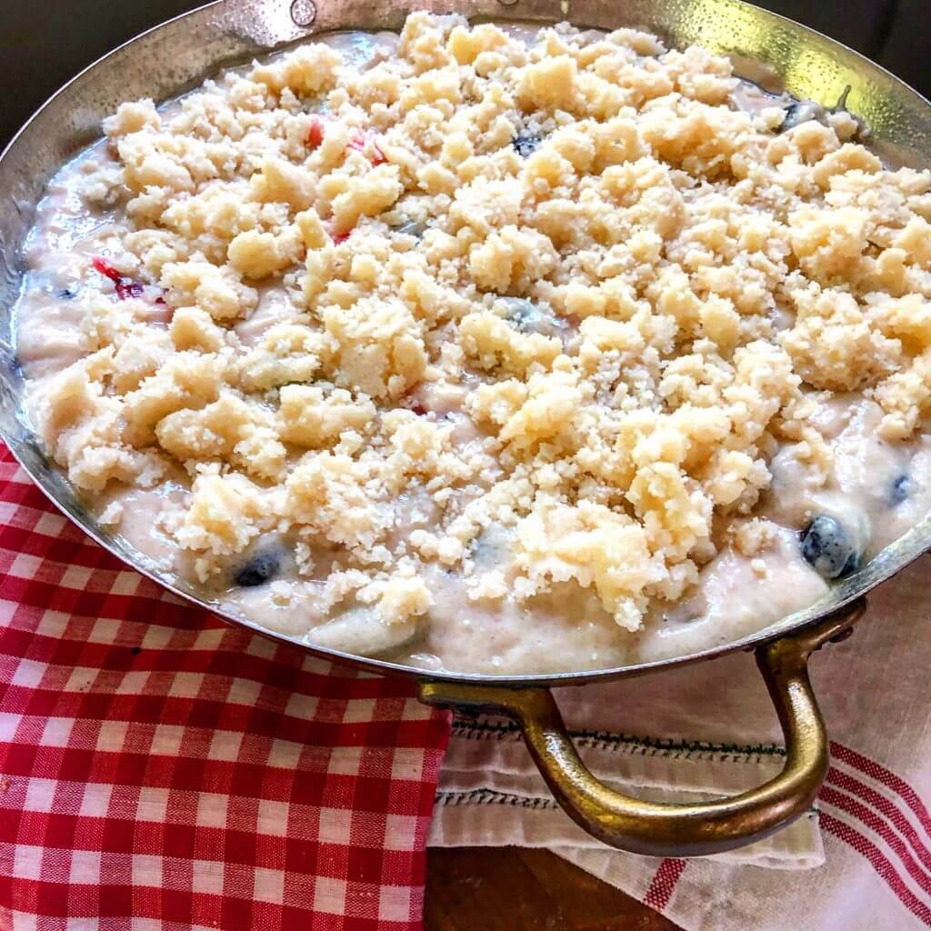 A baking dish filled with berry batter topped with a crumbly streusel mixture, placed on a red and white checkered cloth.