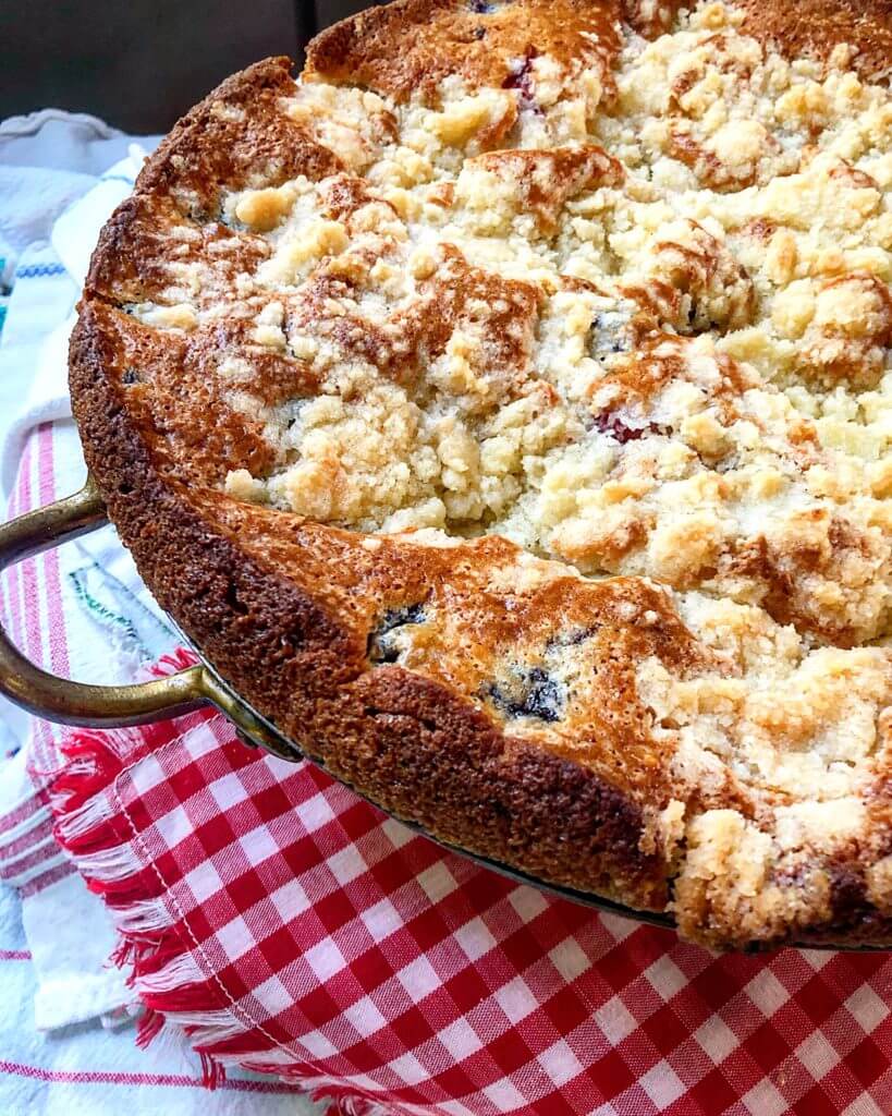 A golden-brown berry crumble cake in a baking dish sits on a red and white checkered cloth.