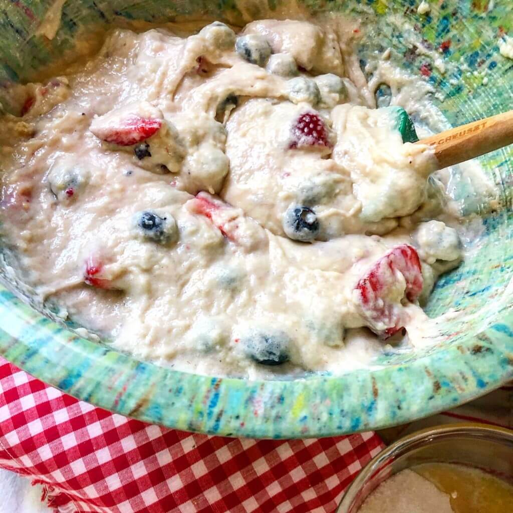 A bowl of mixed berry batter being stirred with a wooden spoon, placed on a red and white checkered cloth.
