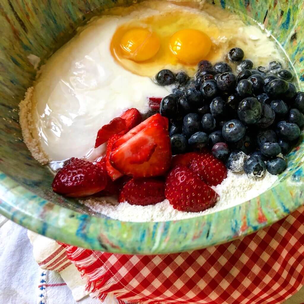 A bowl containing eggs, yogurt, blueberries, and sliced strawberries on top of flour, ready to be mixed, placed on a red and white checkered cloth.