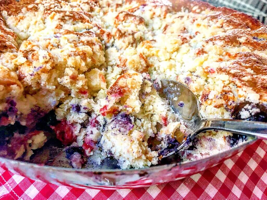 A spoon scooping out a portion of mixed berry crumble cake from a baking dish on a red and white checkered cloth.