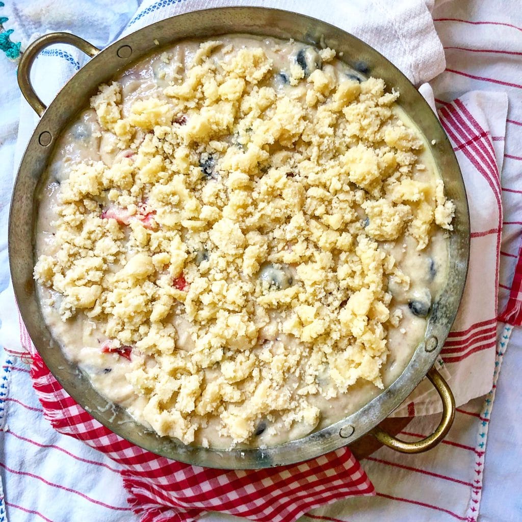 A baking dish filled with berry batter topped with a crumbly streusel mixture, placed on a red and white checkered cloth.