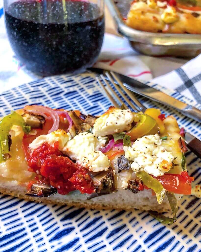 A slice of vegetable and cheese pizza on a blue and white patterned plate, with a glass of red wine in the background.