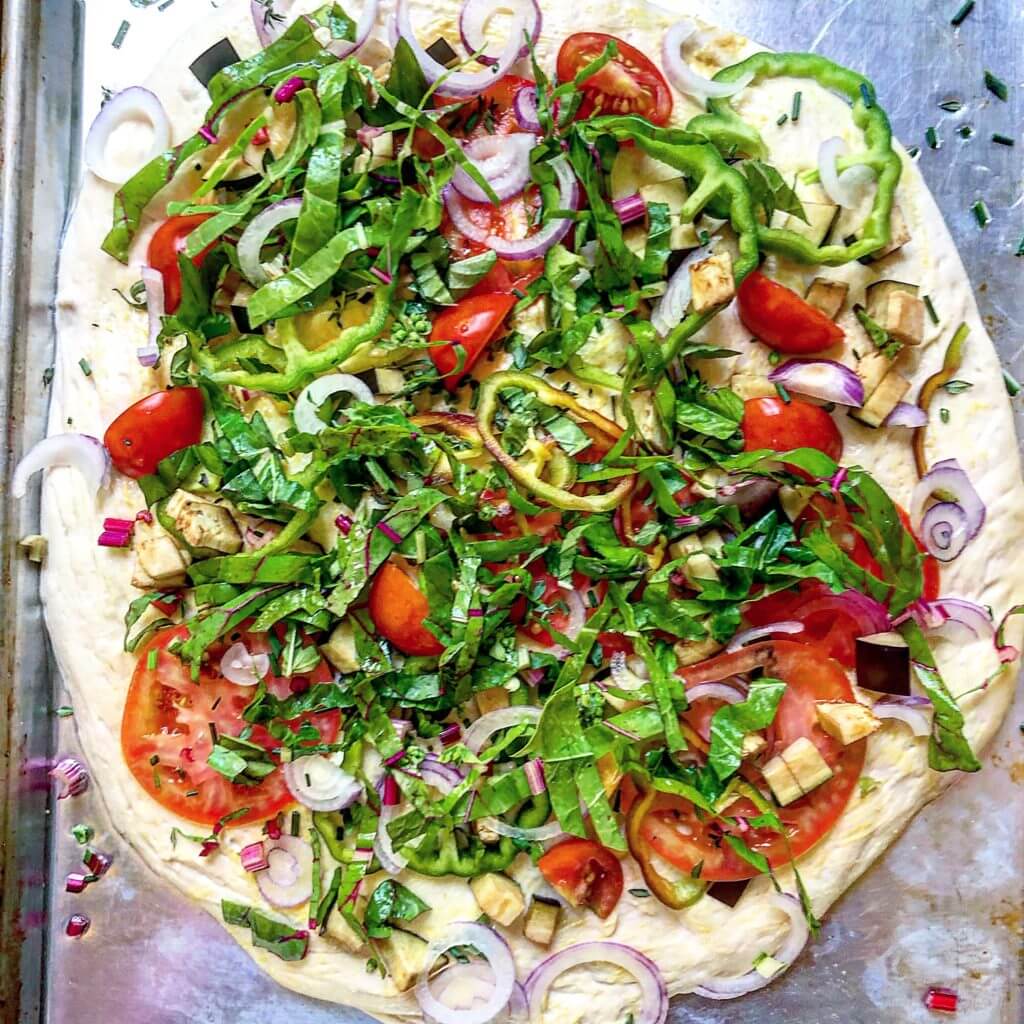 A close-up of an uncooked pizza topped with fresh vegetables, dollops of tomato sauce, and crumbled cheese on a baking sheet.