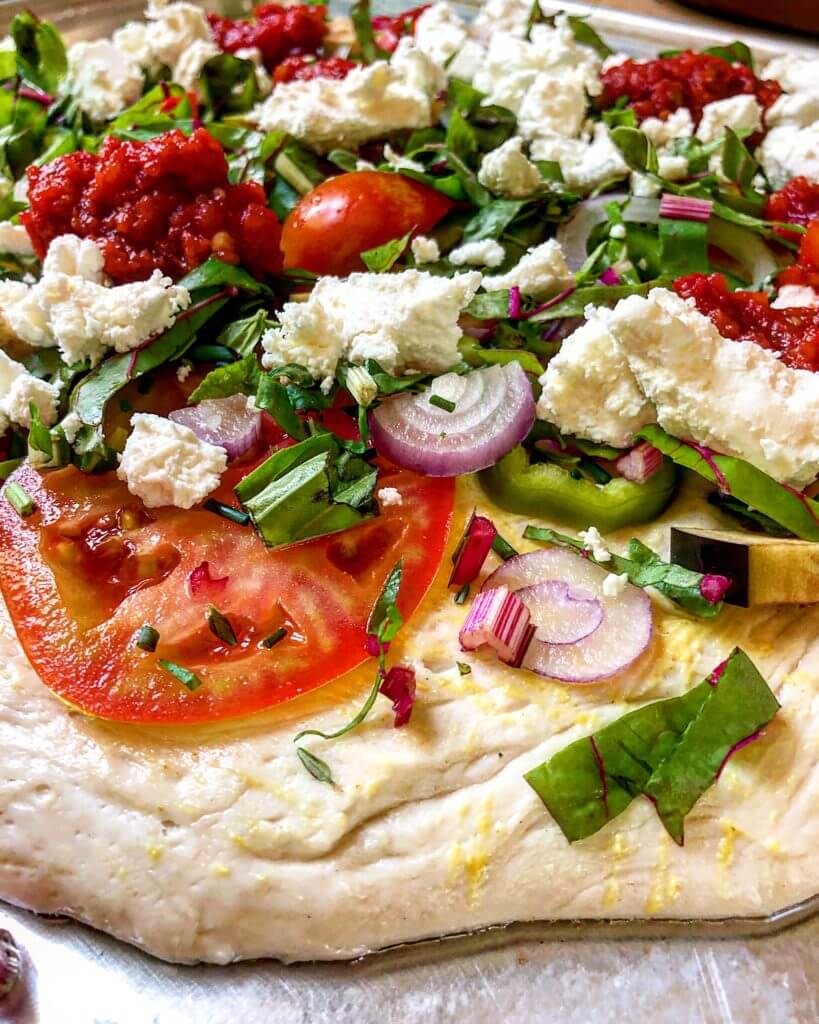 A close-up of an uncooked pizza topped with fresh vegetables, dollops of tomato sauce, and crumbled cheese on a baking sheet.