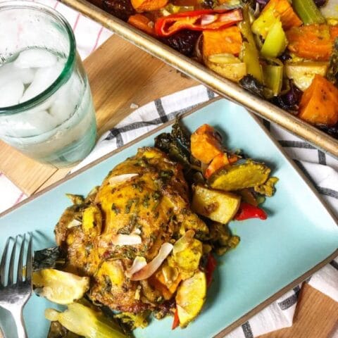 A plate of seasoned chicken thighs and roasted vegetables served next to a glass of iced water, with additional portions in a baking dish nearby.