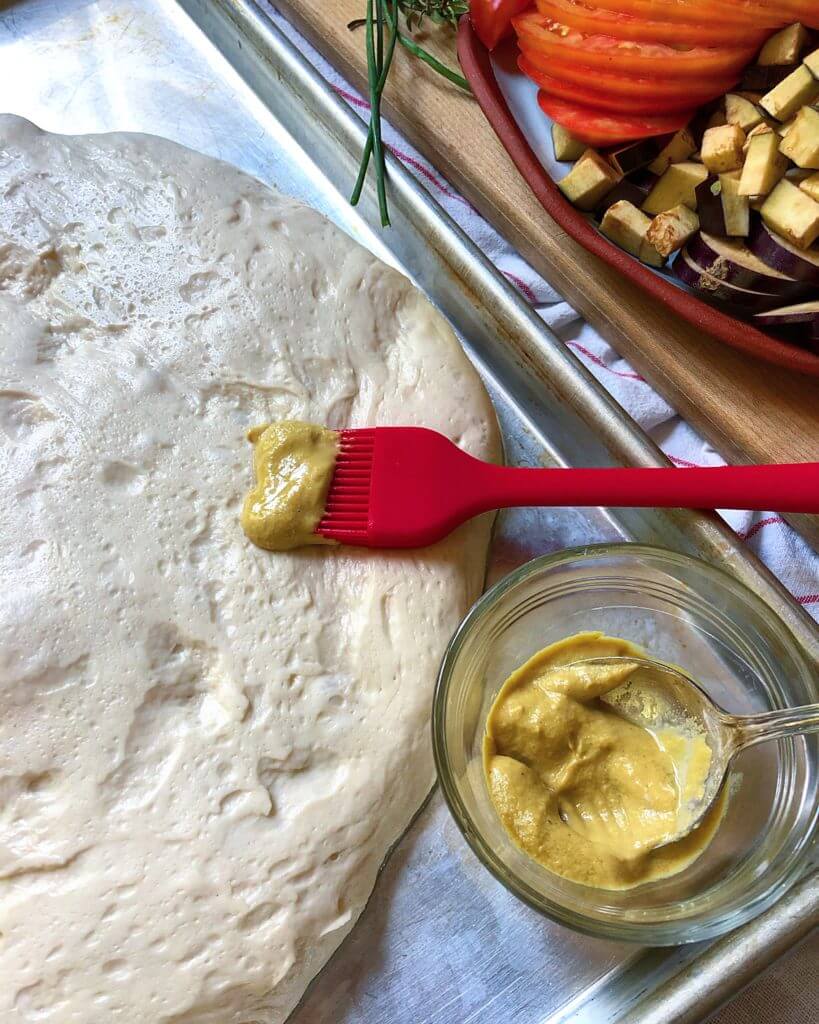 A pizza dough being brushed with mustard using a red brush, with a small glass bowl of mustard and a cutting board with sliced tomatoes and diced eggplant nearby.