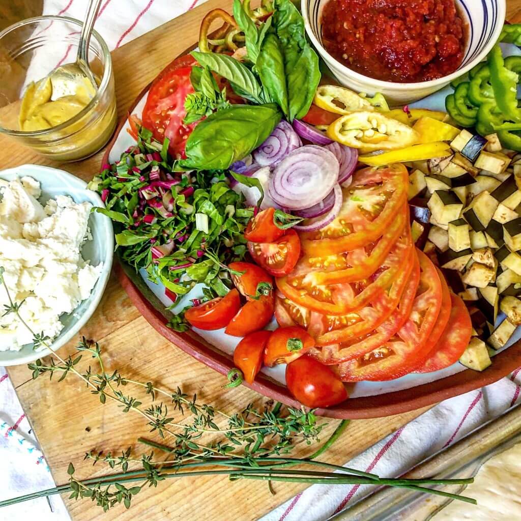 A cutting board with various fresh ingredients, including sliced tomatoes, diced eggplant, chopped greens, sliced onions, basil leaves, and cherry tomatoes, alongside bowls of tomato sauce, mustard, and crumbled cheese.