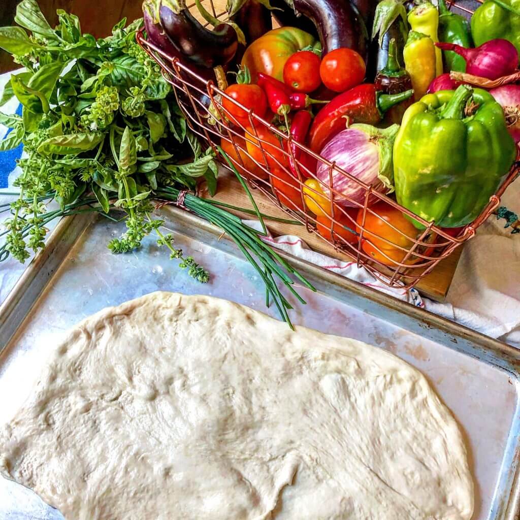 A sheet of rolled-out pizza dough on a baking tray next to a basket filled with fresh vegetables, including bell peppers, tomatoes, eggplants, and onions, with a bundle of fresh herbs on the side.