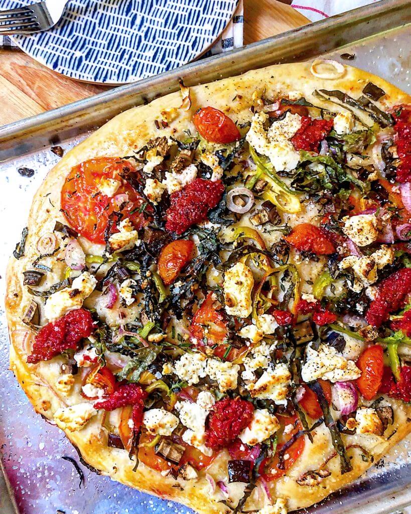 A freshly baked vegetable pizza with a golden crust, topped with tomatoes, onions, greens, dollops of tomato sauce, and crumbled cheese on a baking sheet.