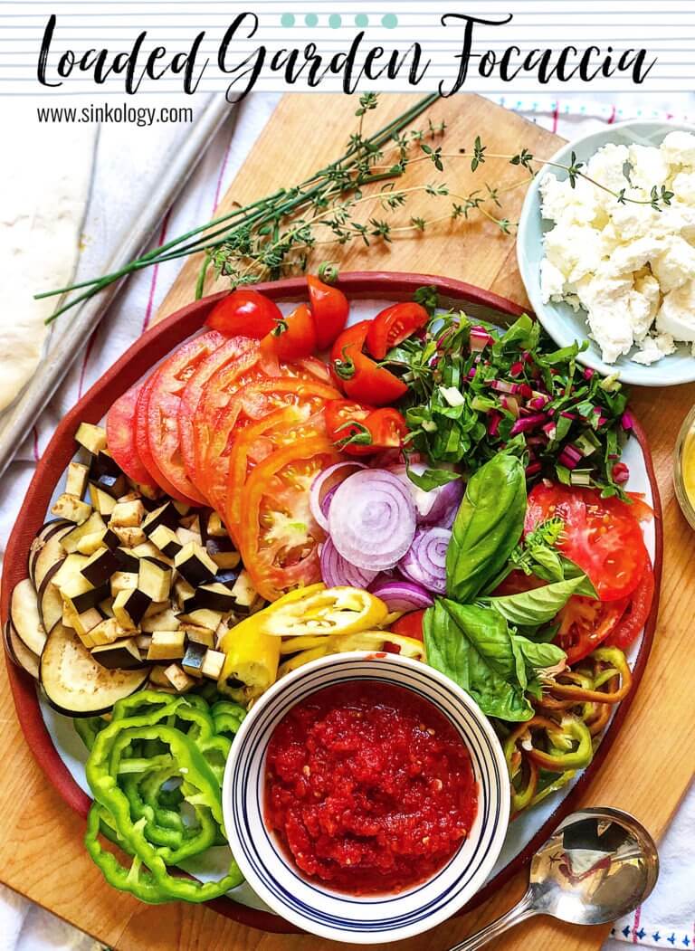A cutting board with various fresh ingredients for a focaccia, including sliced tomatoes, diced eggplant, sliced onions, basil leaves, and a bowl of tomato sauce, labeled "Loaded Garden Focaccia" from sinkology.com.