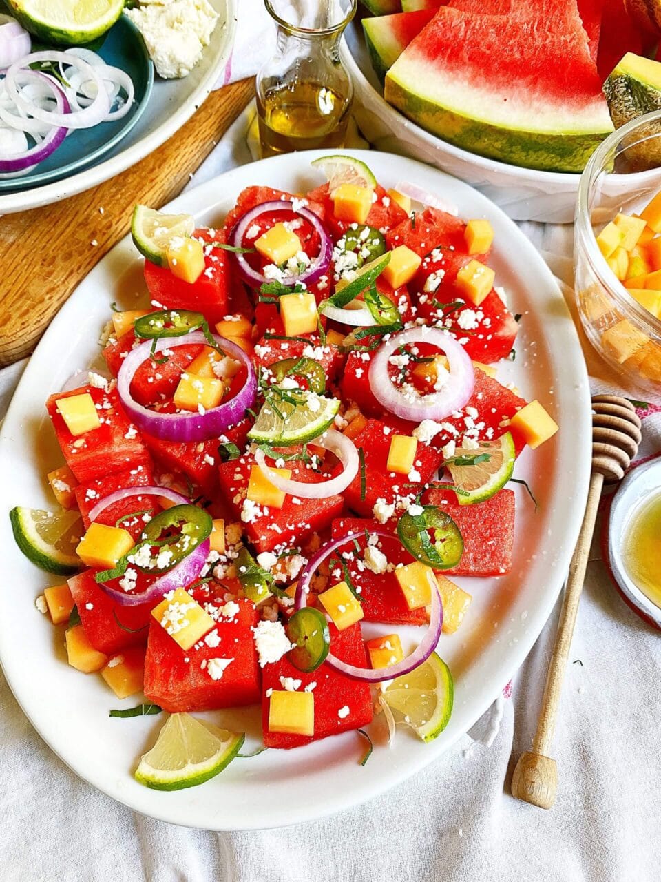 Plate of watermelon salad topped with cheese cubes, red onion rings, jalapeno slices, and lime wedges, surrounded by additional ingredients and dressing.
