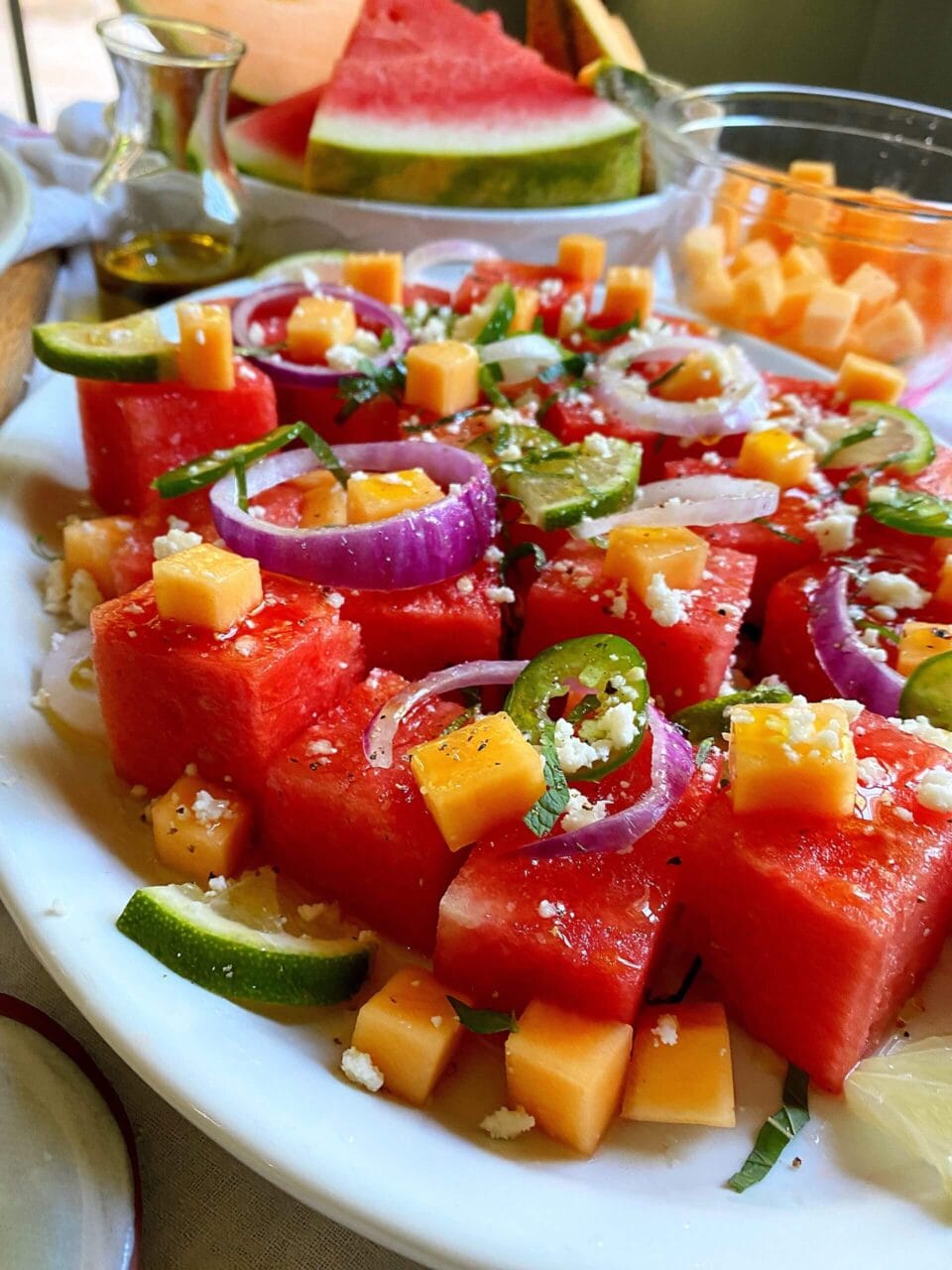 Plate of watermelon cubes topped with cheese, red onion, and jalapeno slices, with additional fruit and a small bottle of dressing in the background.