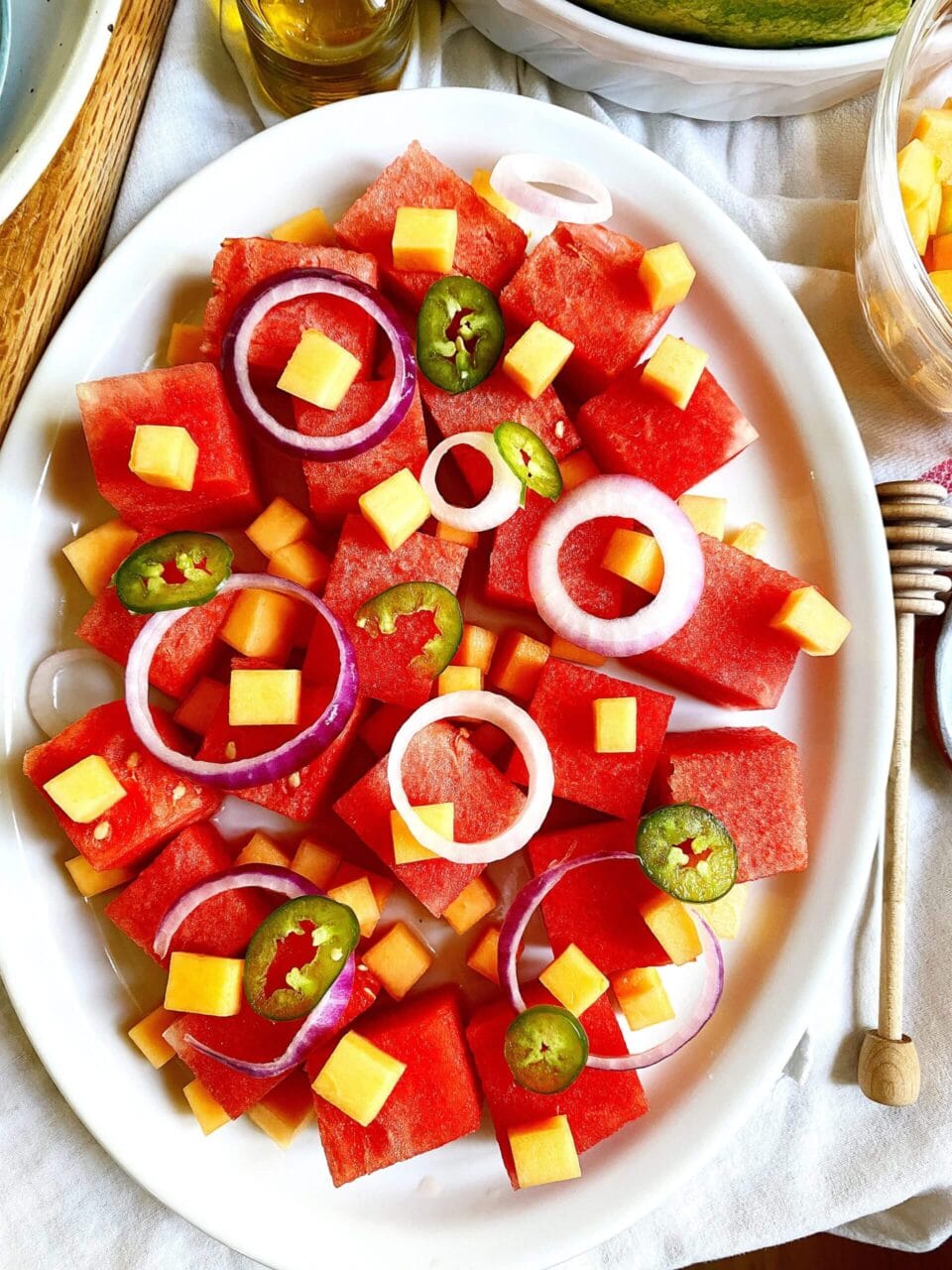 Close-up of a watermelon salad with cheese cubes, red onion rings, and jalapeno slices, garnished with a small bottle of dressing on the side.