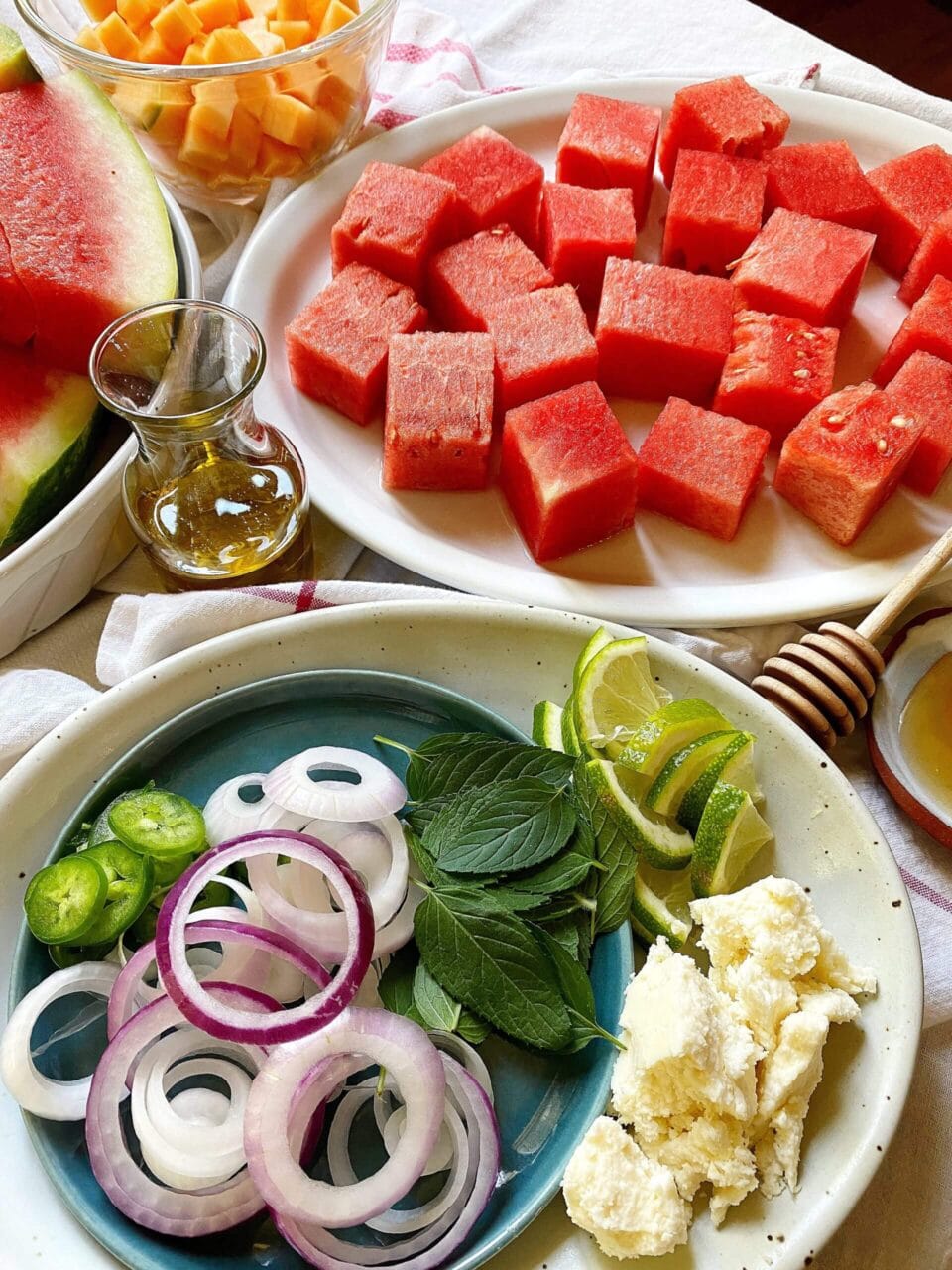 Ingredients for a watermelon salad, including watermelon cubes, sliced onions, mint leaves, lime wedges, cheese, and a small bottle of dressing.