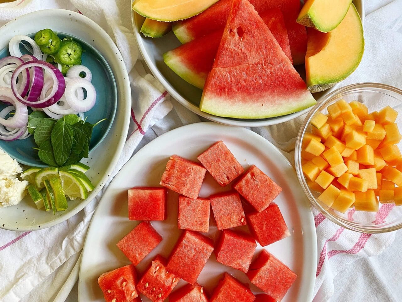Ingredients for a watermelon salad, including watermelon cubes, sliced onions, mint leaves, lime wedges, cheese, cantaloupe slices, and a bowl of cheese cubes.